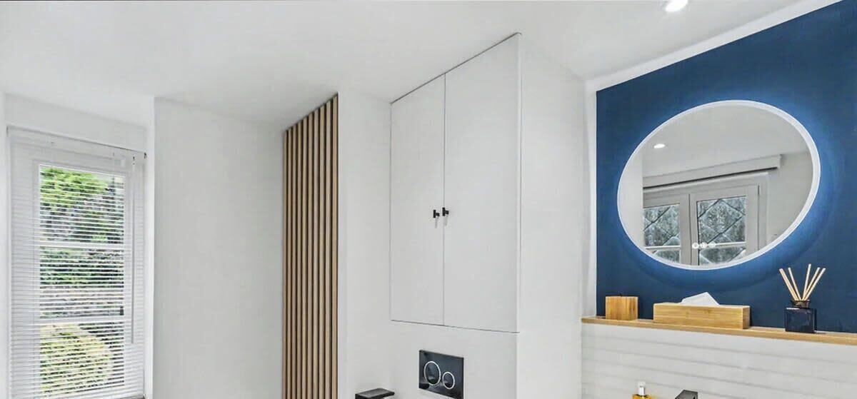 Un baño con un armario blanco, un revestimiento de listones de madera, un espejo redondo y un tocador de madera con lavabo.