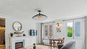 A living room with a fireplace, a dining area with a table and chairs, a ceiling fan, and a large window with a view. - La petite source: modern apartment in the heart of Honfleur (Honfleur)