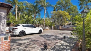 A white car parked in a driveway with a basketball hoop, palm trees, and a brick wall.