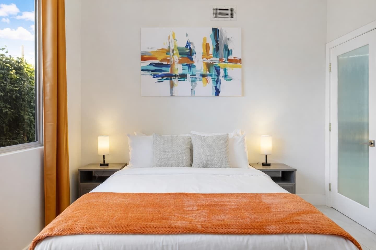 Une chambre d’hôtel avec un lit, deux tables de chevet avec des lampes, une fenêtre avec des rideaux et un tableau au mur.