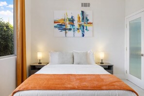 Una habitación de hotel con una cama, dos mesitas de noche con lámparas, una ventana con cortinas y un cuadro en la pared.