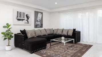 A modern living room with a black sectional sofa, patterned throw pillows, a glass coffee table, and a large potted plant.