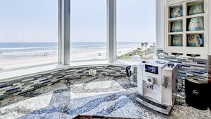 Ett modernt badrum med ett stort fönster med utsikt över en strand, en kaffemaskin och en kakelbeklädd vägg bakom handfatet.