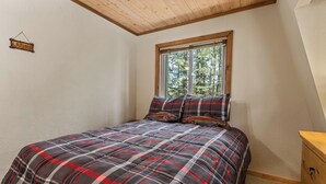 A bedroom with a plaid bedspread, a window with a view of trees, a wooden ceiling, and a sign on the wall. - Summit Mountain Lodge (Big Bear Lake)