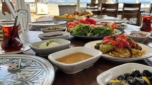 A table set for a meal with various dishes, including a bowl of olives, a plate of vegetables, and a glass of tea, overlooking a beach. - Apelles Butik Otel (Seferihisar)