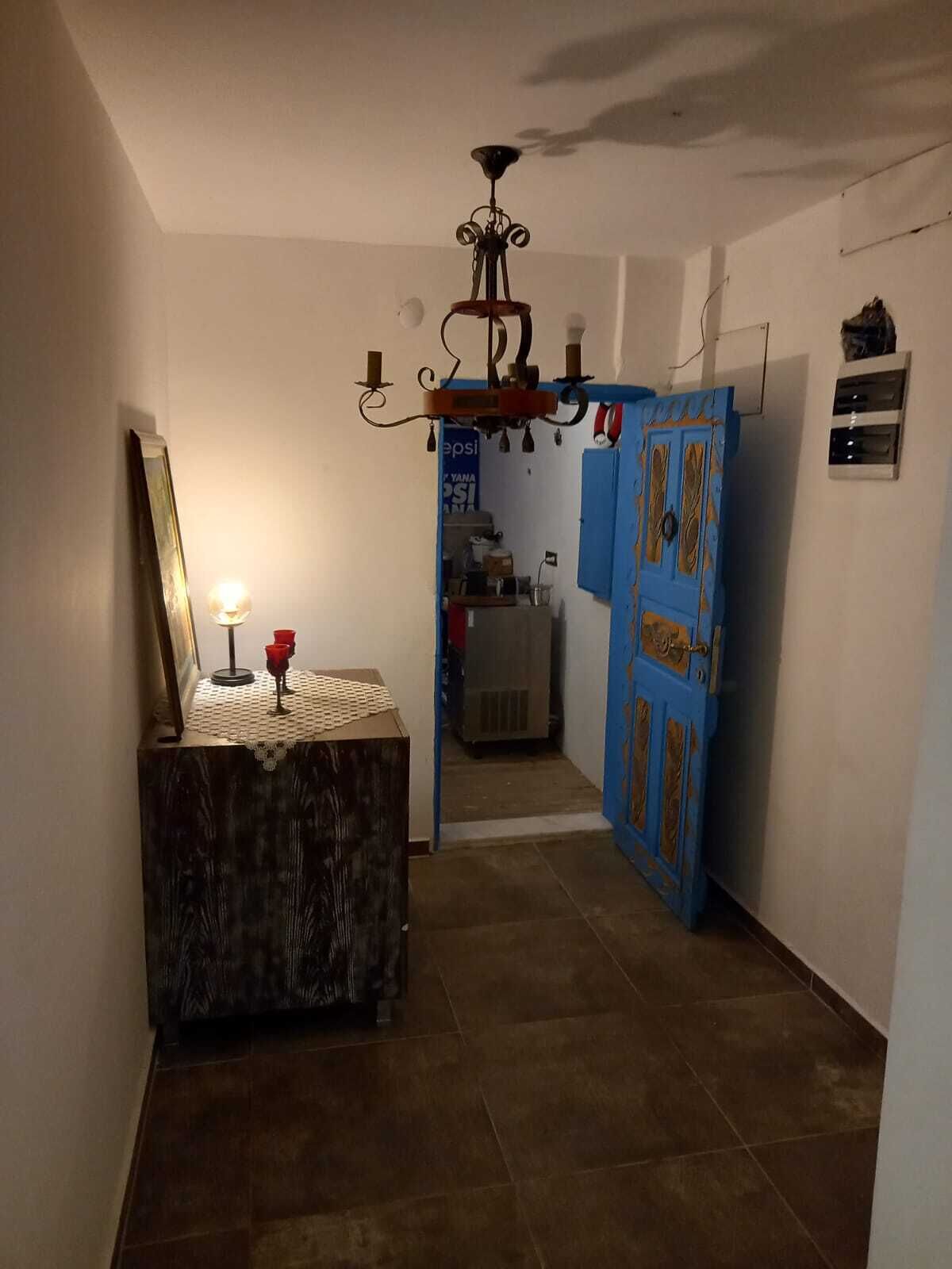Passadís amb un armari de fusta, un mirall i una làmpada encèsa. Hi ha una porta blava amb panells decoratius i un ventilador de sostre.