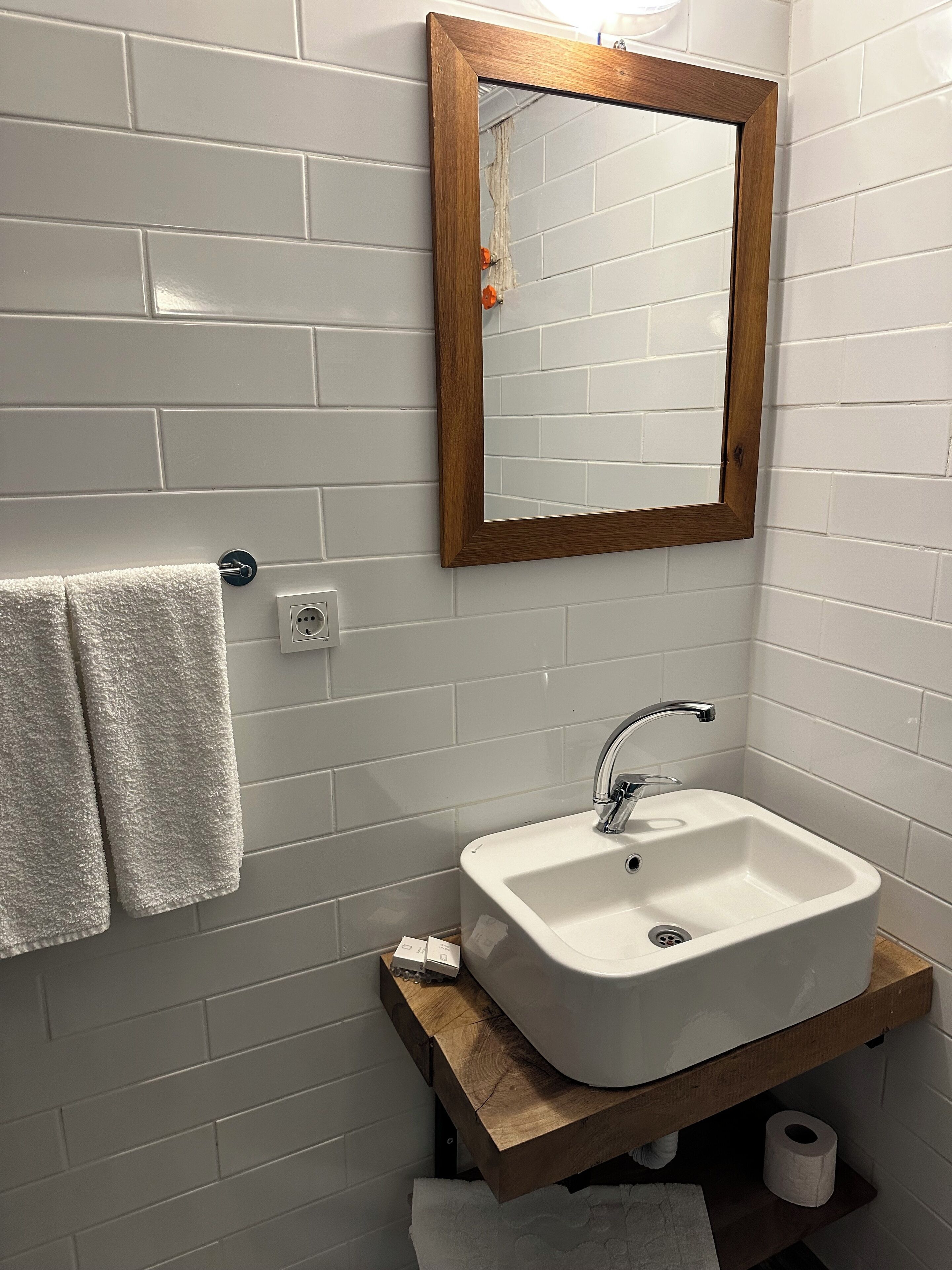 Une salle de bain avec des murs recouverts de carreaux blancs, un miroir encadré en bois, un lavabo blanc avec un robinet chromé et une serviette suspendue à un porte-serviettes.