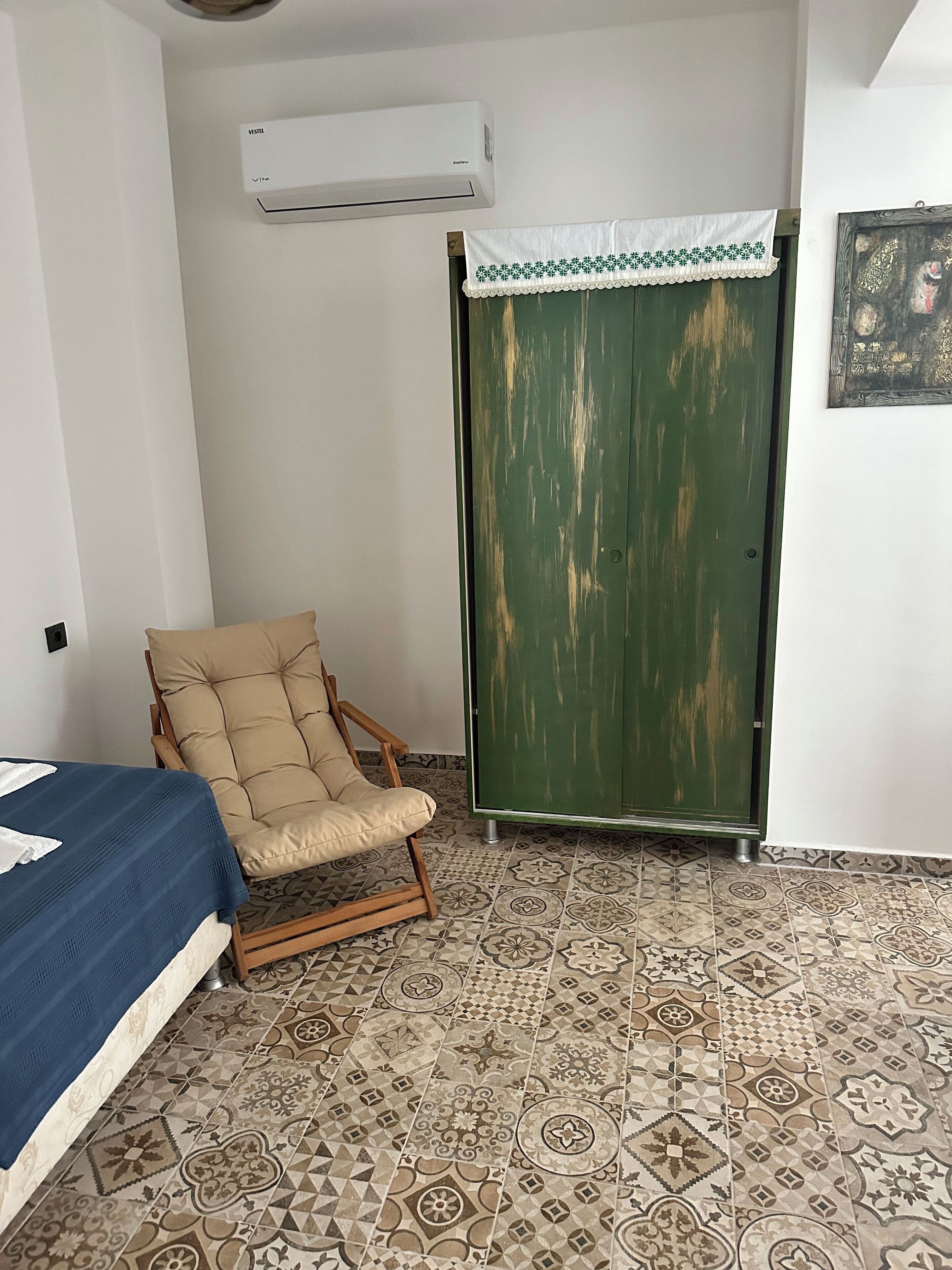 Una habitación con cama, una silla, un armario verde, baldosas de suelo con diseño y un equipo de aire acondicionado.
