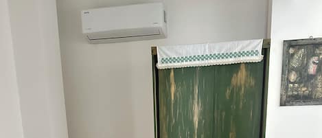Una habitación con cama, una silla, un armario verde, baldosas de suelo con diseño y un equipo de aire acondicionado.