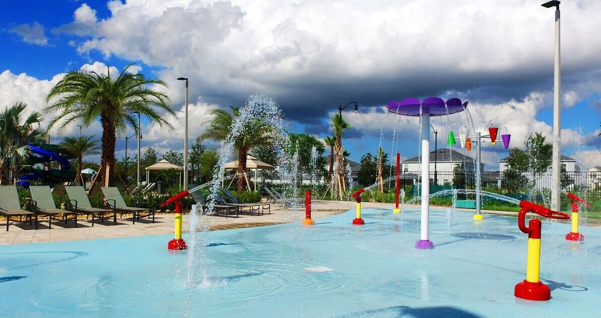 A water park with multiple water features, including fountains and water slides, surrounded by palm trees and lounge chairs.