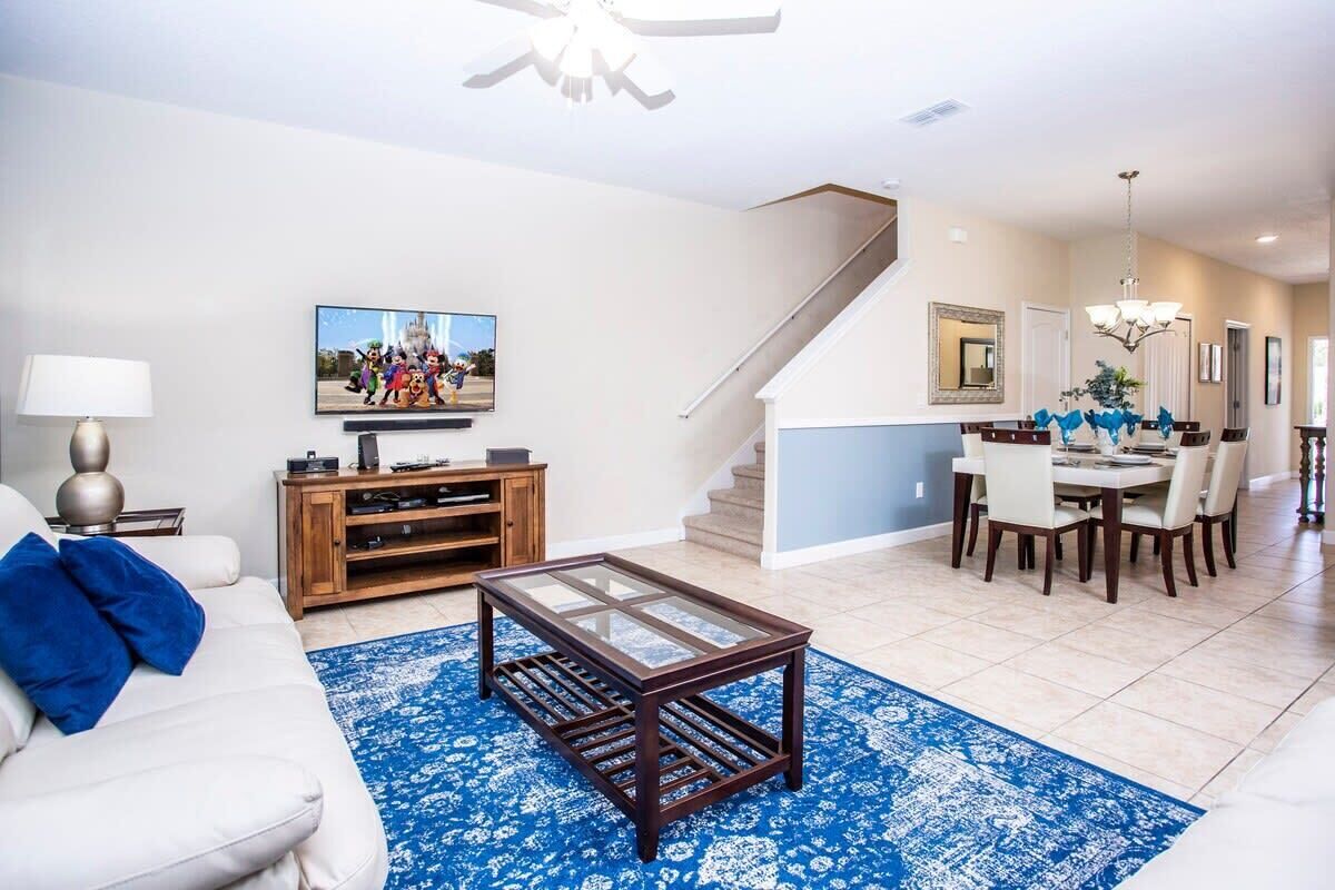 A spacious living room with a white sofa, a coffee table, and a blue patterned rug. A dining area with a chandelier and a staircase is visible in the background.