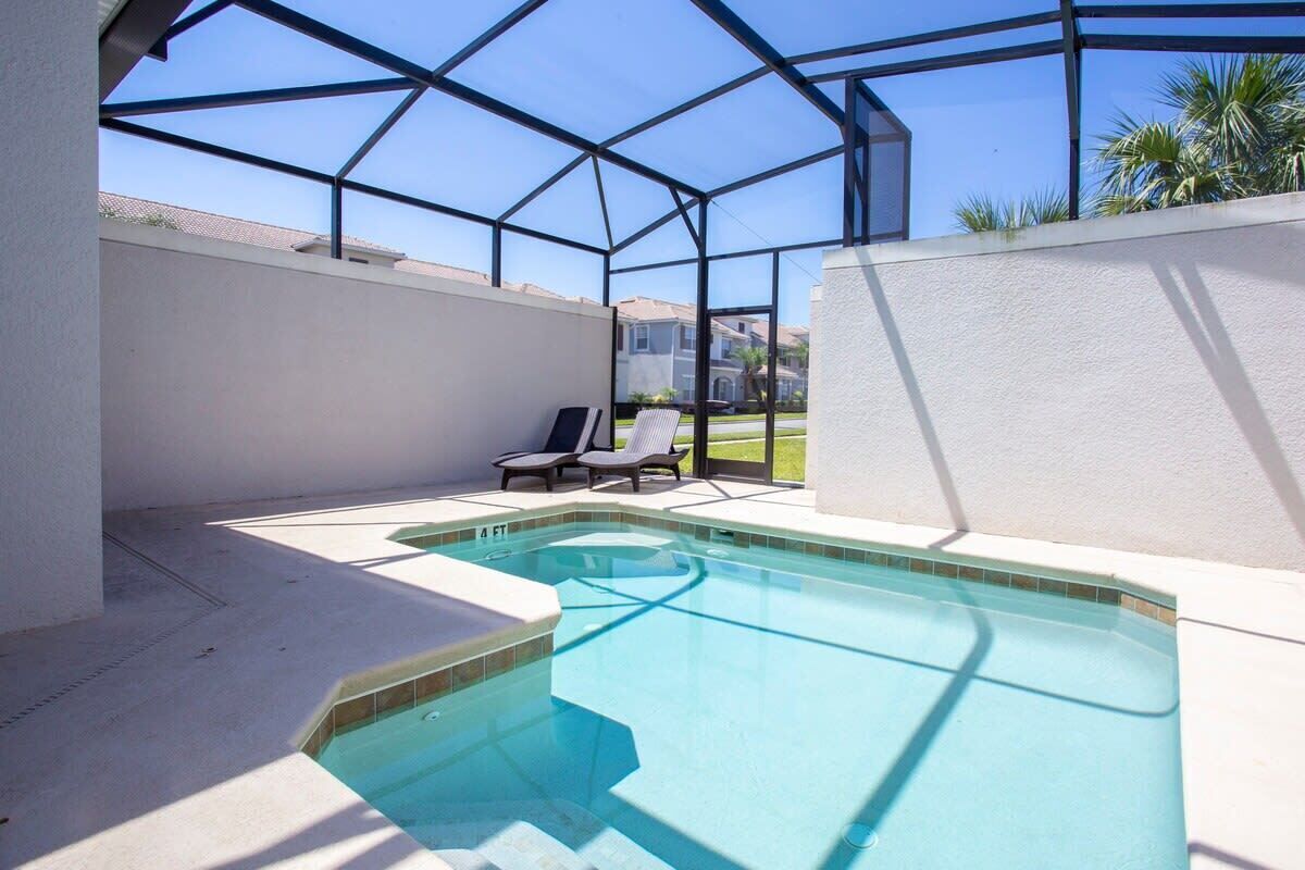 A swimming pool with a glass enclosure, two lounge chairs, and a view of a residential area.