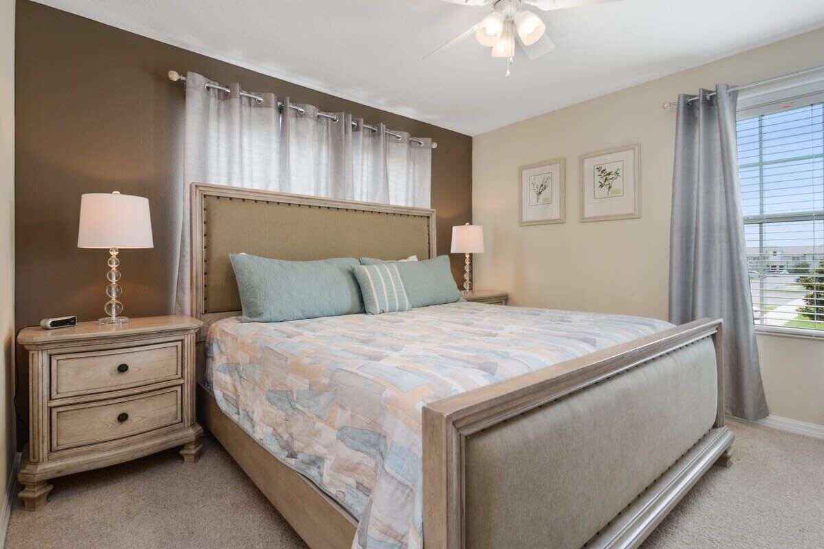A bedroom with a large bed, two bedside tables with lamps, a ceiling fan, and a window with curtains.