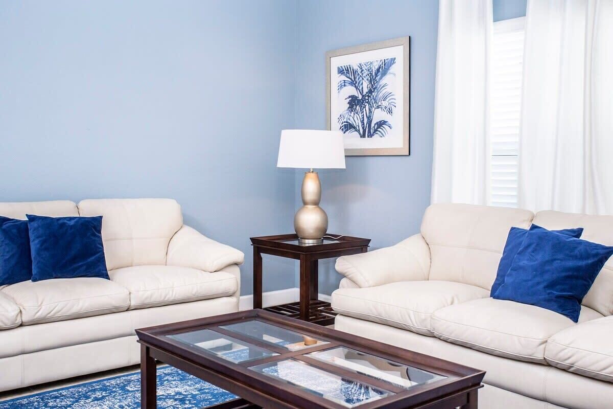 A living room with two cream sofas, a glass coffee table, a small wooden side table with a lamp, and a framed picture on the wall.
