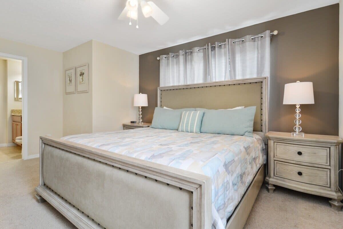A bedroom with a large bed, two bedside tables with lamps, a ceiling fan, and a window with curtains.
