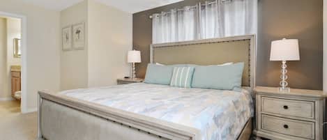A bedroom with a large bed, two bedside tables with lamps, a ceiling fan, and a window with curtains.