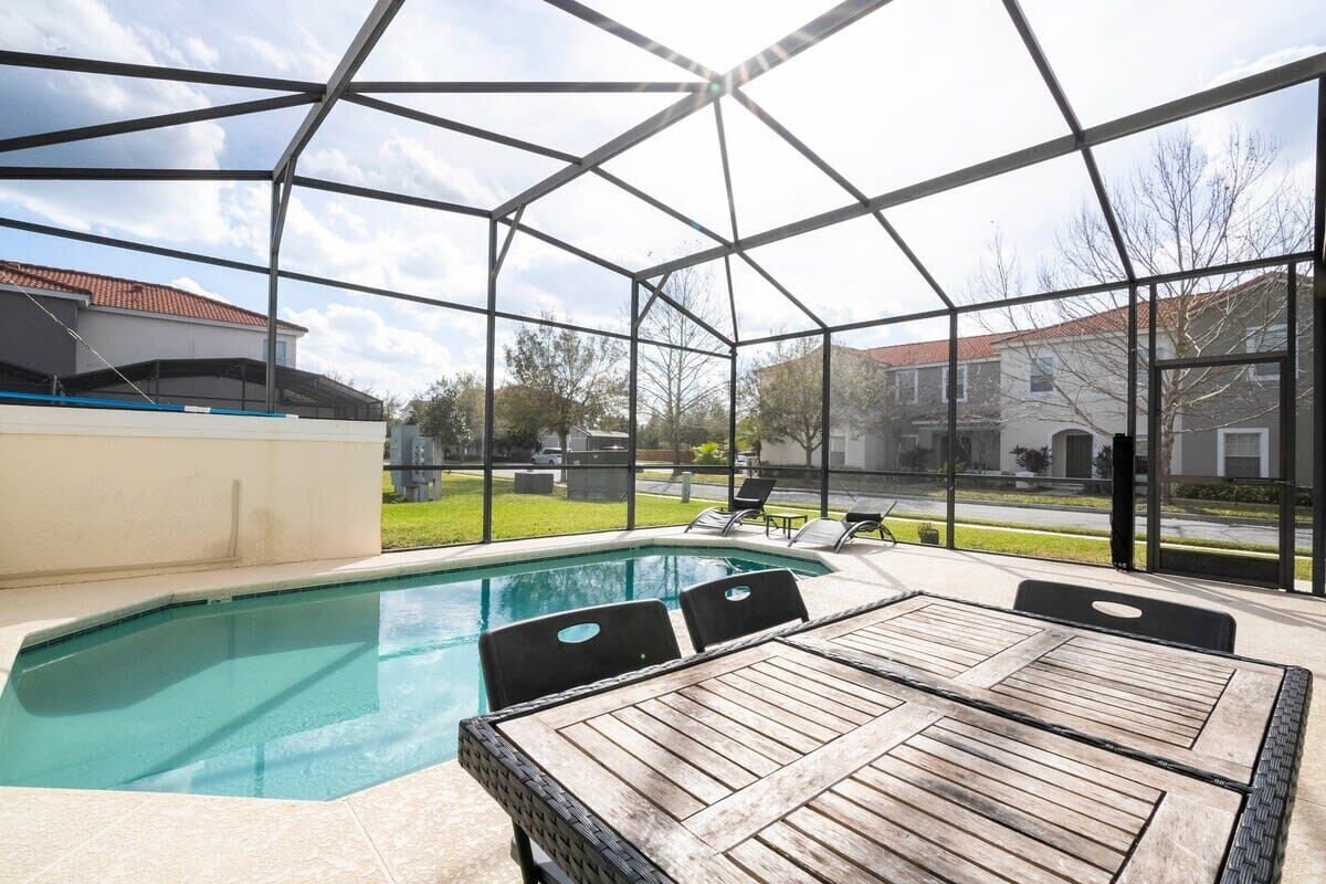 A covered outdoor swimming pool with a wooden table and chairs, surrounded by a metal frame structure.
