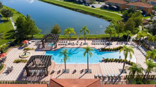 An aerial view of a resort with a central swimming pool, surrounded by lounge chairs and palm trees, with a body of water and buildings in the background. - Amazing 4Bd Screened Pool Close to Disney 4572 (Kissimmee)
