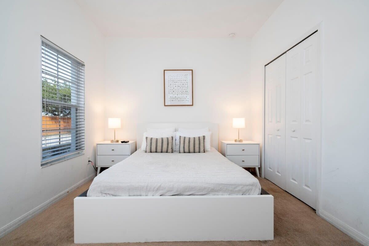 A bedroom with a bed, two bedside tables, a window with blinds, a framed picture on the wall, and a wardrobe.