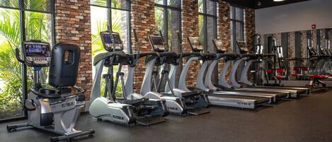 A gym with multiple treadmills lined up against a brick wall with large windows.