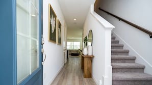 A hallway with a blue door, framed artwork, a wooden console table with vases, and a carpeted staircase. - Casa Serene 3Bd near Disney @Festival Resort 963 (Davenport)