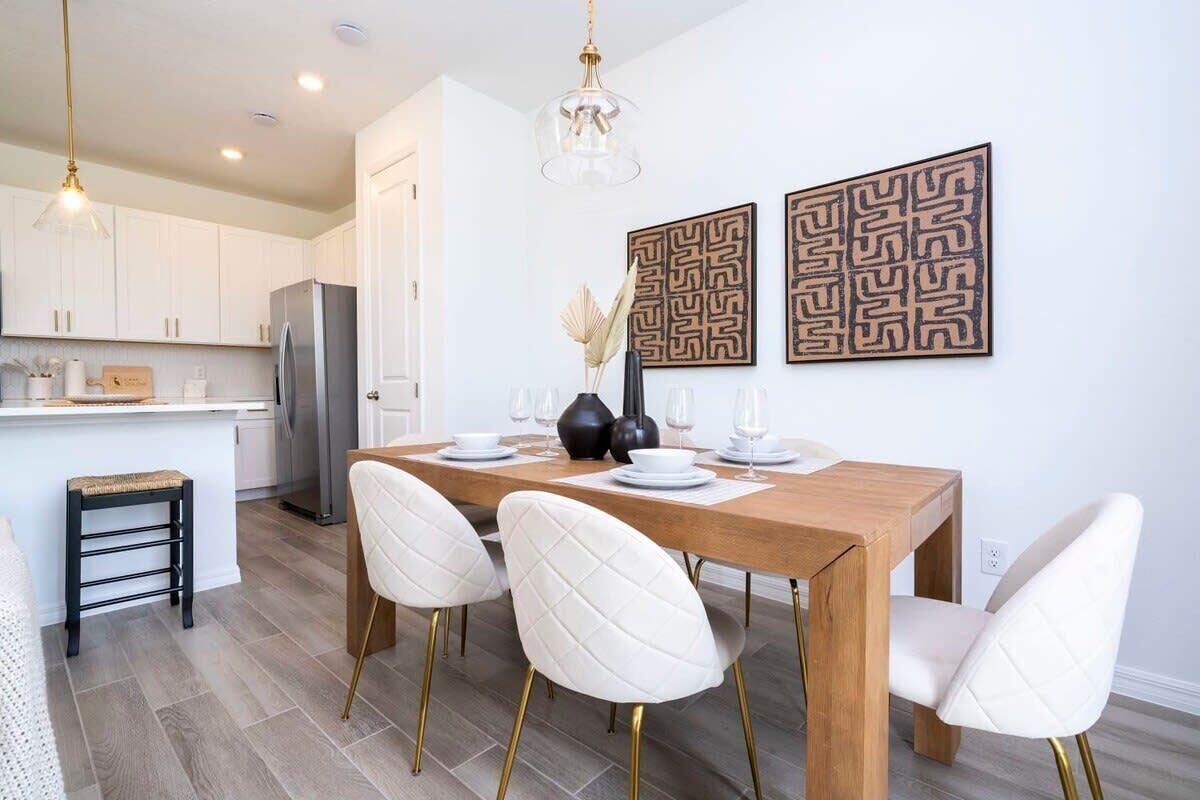 A modern kitchen with white cabinets, a stainless steel refrigerator, and a wooden dining table with white chairs. There are two wall-mounted light fixtures and decorative wall art.