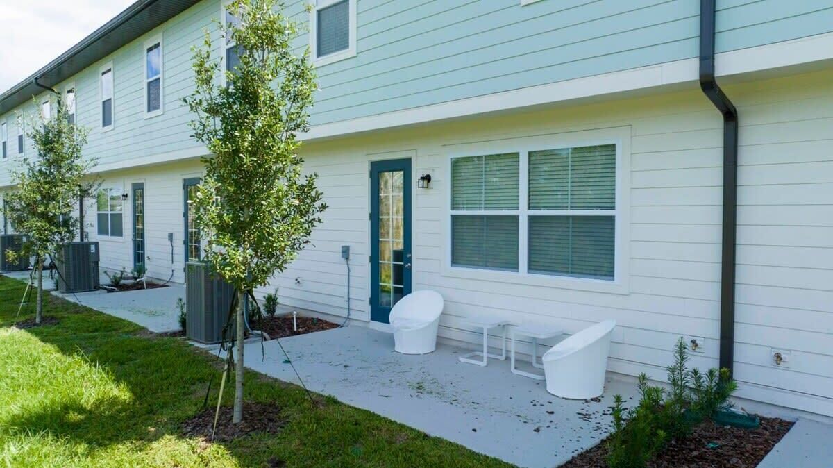 A modern apartment building with a light blue exterior, a covered outdoor seating area, and a small garden.