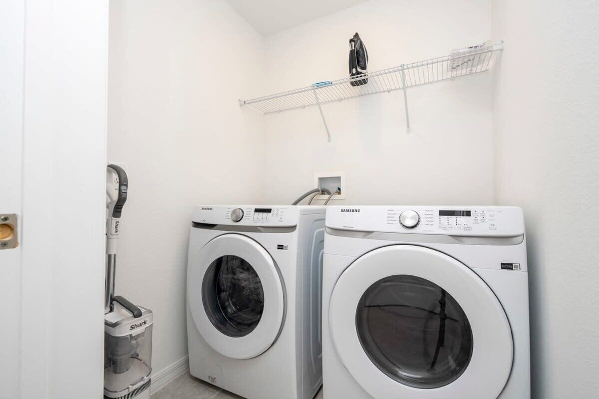 A laundry room with two front-loading washing machines, a vacuum cleaner, and a metal shelf with a hanging iron.