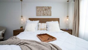 Een net opgemaakt bed met een houten dienblad, wit beddengoed en een bloemrijk kunstwerk aan de muur.