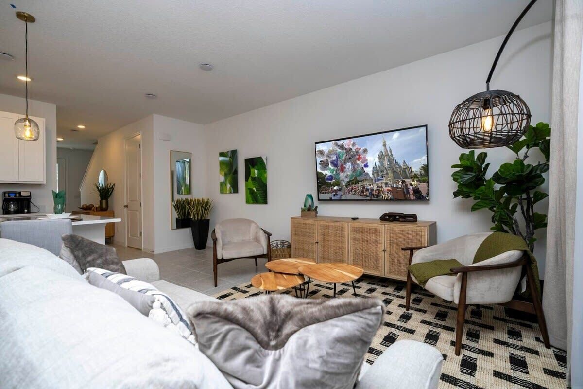 Ein modernes Wohnzimmer mit einer Couch, Sesseln, einem Couchtisch und einem Fernseher. Im Hintergrund ist eine Küche zu sehen und eine Treppe führt zu einer weiteren Ebene.