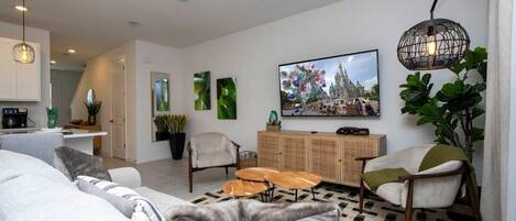 Ein modernes Wohnzimmer mit einer Couch, Sesseln, einem Couchtisch und einem Fernseher. Im Hintergrund ist eine Küche zu sehen und eine Treppe führt zu einer weiteren Ebene.