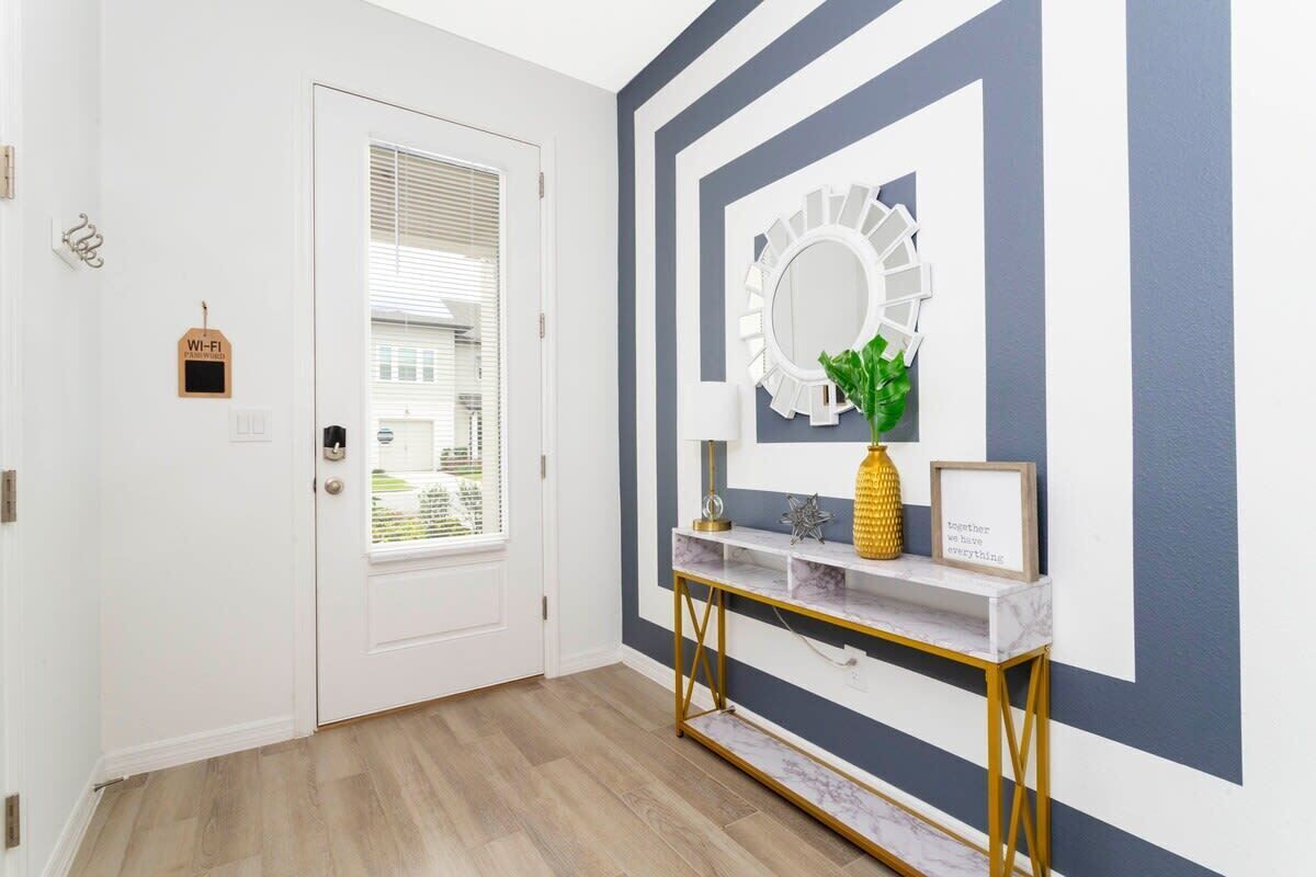 A hallway with a white door, a wall-mounted mirror, a gold console table with a pineapple decoration, and a framed inspirational quote.