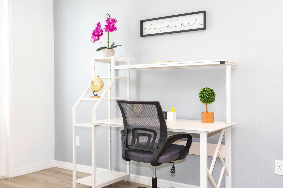 A white desk with a mesh chair, a small potted plant, and a framed sign on the wall.