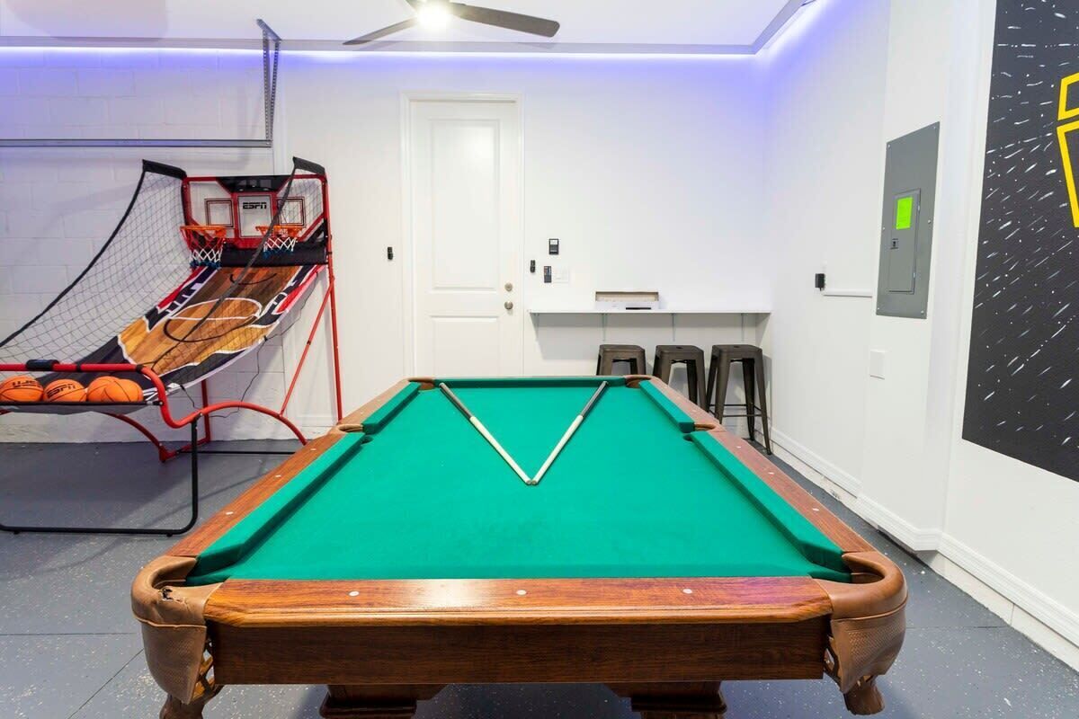 A pool table with a green felt surface, a wooden frame, and a corner pocket. There is a white door and a wall-mounted shelf with items on it.
