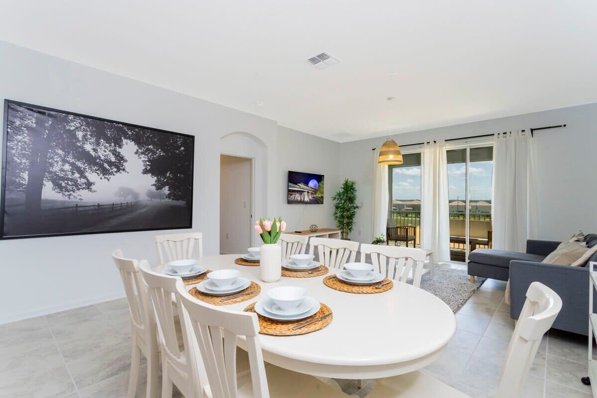 A dining area with a round table set for four, surrounded by white chairs. There is a large framed picture on the wall, a potted plant, and a view of the outdoors through large windows.