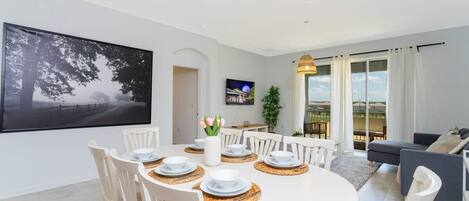 A dining area with a round table set for four, surrounded by white chairs. There is a large framed picture on the wall, a potted plant, and a view of the outdoors through large windows.