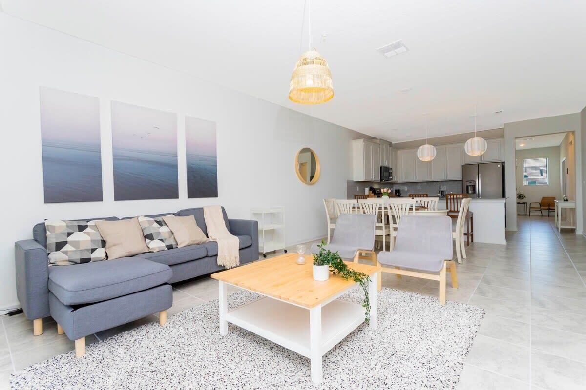 A modern living room with a grey sofa, a wooden coffee table, and a dining area with white chairs.