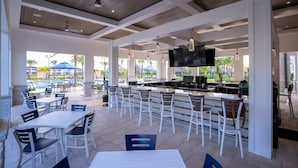 Un espace bar extérieur avec des tables et des chaises blanches, un téléviseur à écran plat fixé au-dessus du bar, et une piscine visible au fond.