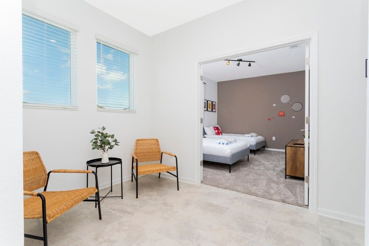 A modern, well-lit room with a large window, two wicker chairs, a small table with a plant, and a bed with a white mattress and a gray headboard.