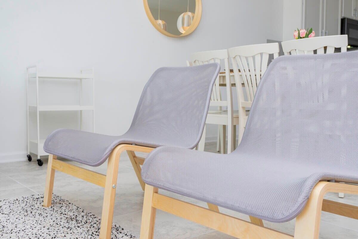 A room with two wooden chairs with mesh backs and seats, a white table, a white shelf unit, and a black and white speckled rug.