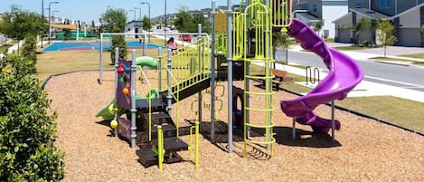 Un parque infantil con un tobogán y estructuras para escalar, rodeado por una calle y edificios residenciales.