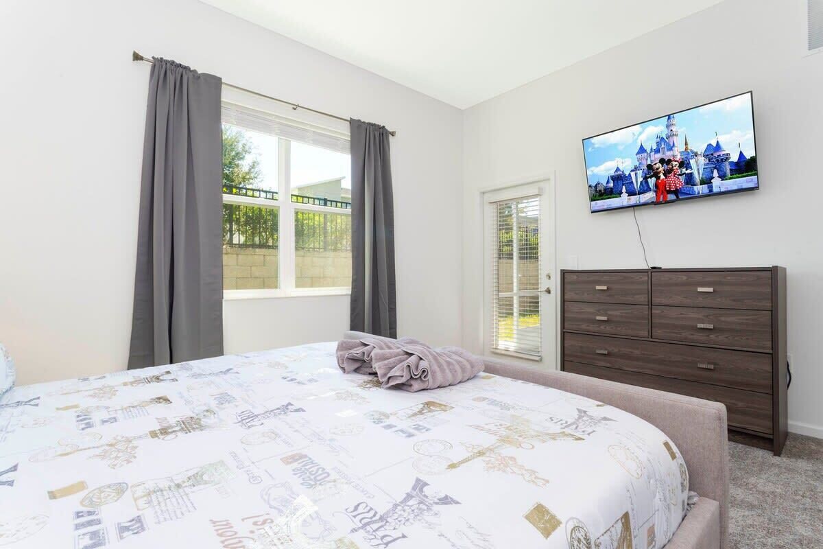 A bedroom with a bed, a TV mounted on the wall, a dresser, and a window with curtains.