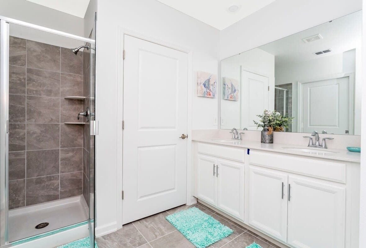 Un bagno moderno con doccia vetrata, mobili bianchi, uno specchio grande e un tappeto color petrolio.