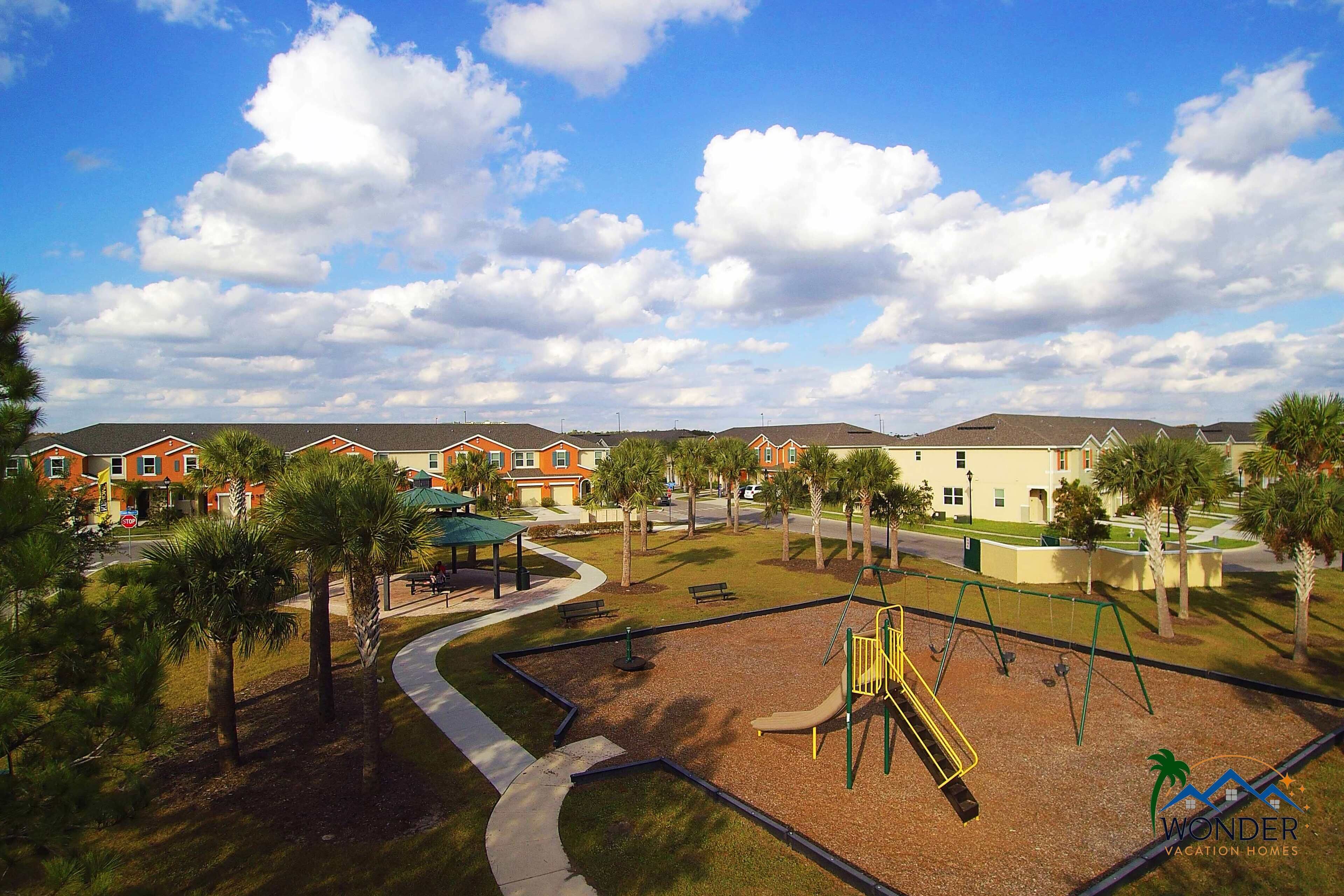 A playground with swings and a slide, surrounded by palm trees and a walking path.