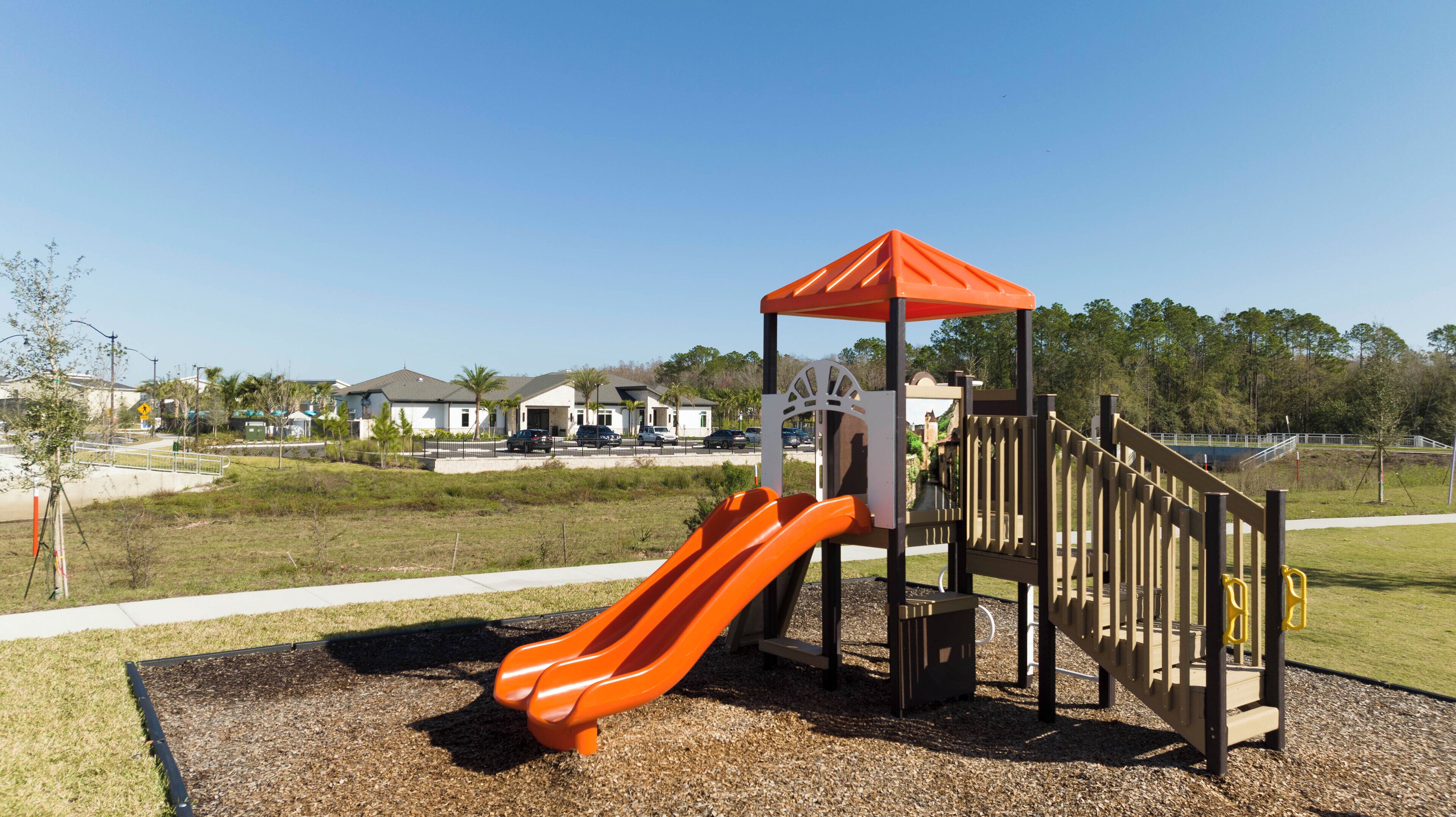 A playground with a slide and climbing structure, surrounded by a grassy area and residential buildings in the background.