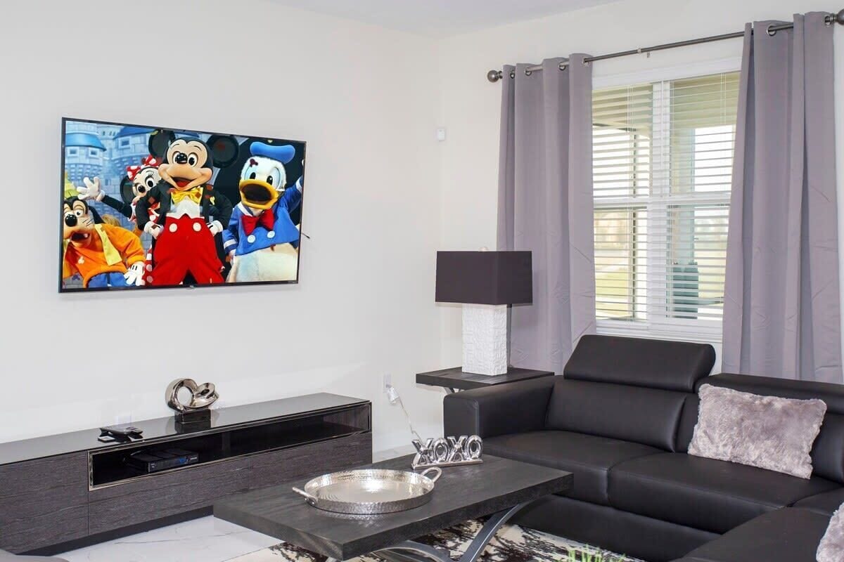 A living room with a large flat-screen TV displaying animated characters, a black leather sofa, a side table with a lamp, and a window with curtains.