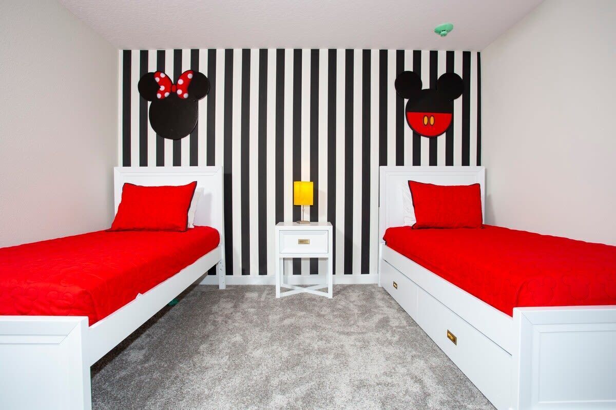 A room with two beds, each with a red bedspread and white headboards. There is a small white nightstand with a lamp between the beds. The wall behind the beds has a black and white striped pattern with two large Mickey Mouse head silhouettes.