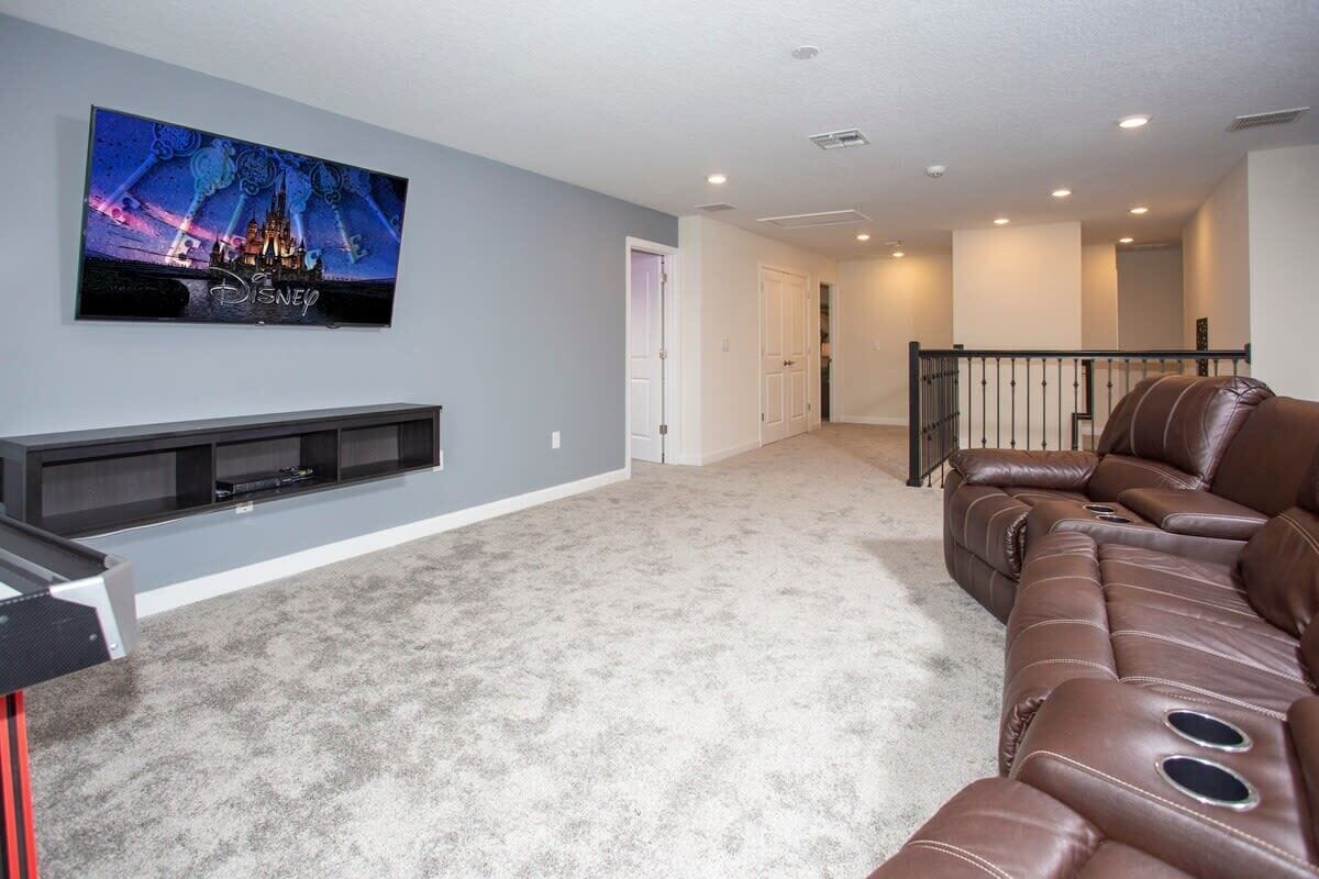 A living room with a flat-screen TV mounted on the wall, a brown leather sofa, and a carpeted floor.