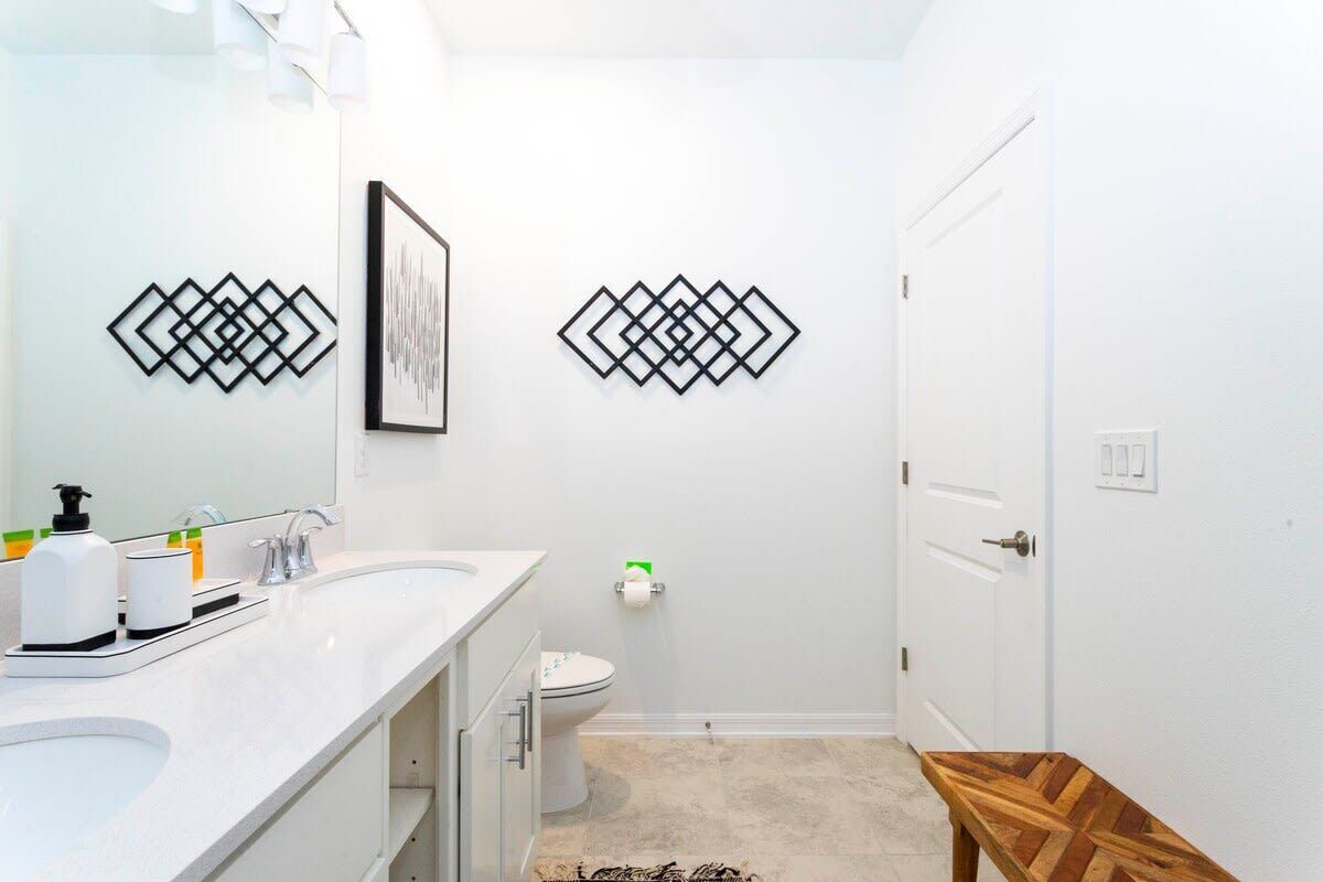 A modern bathroom with a white sink, mirror, and toilet. There are decorative wall art pieces and a wooden bench.
