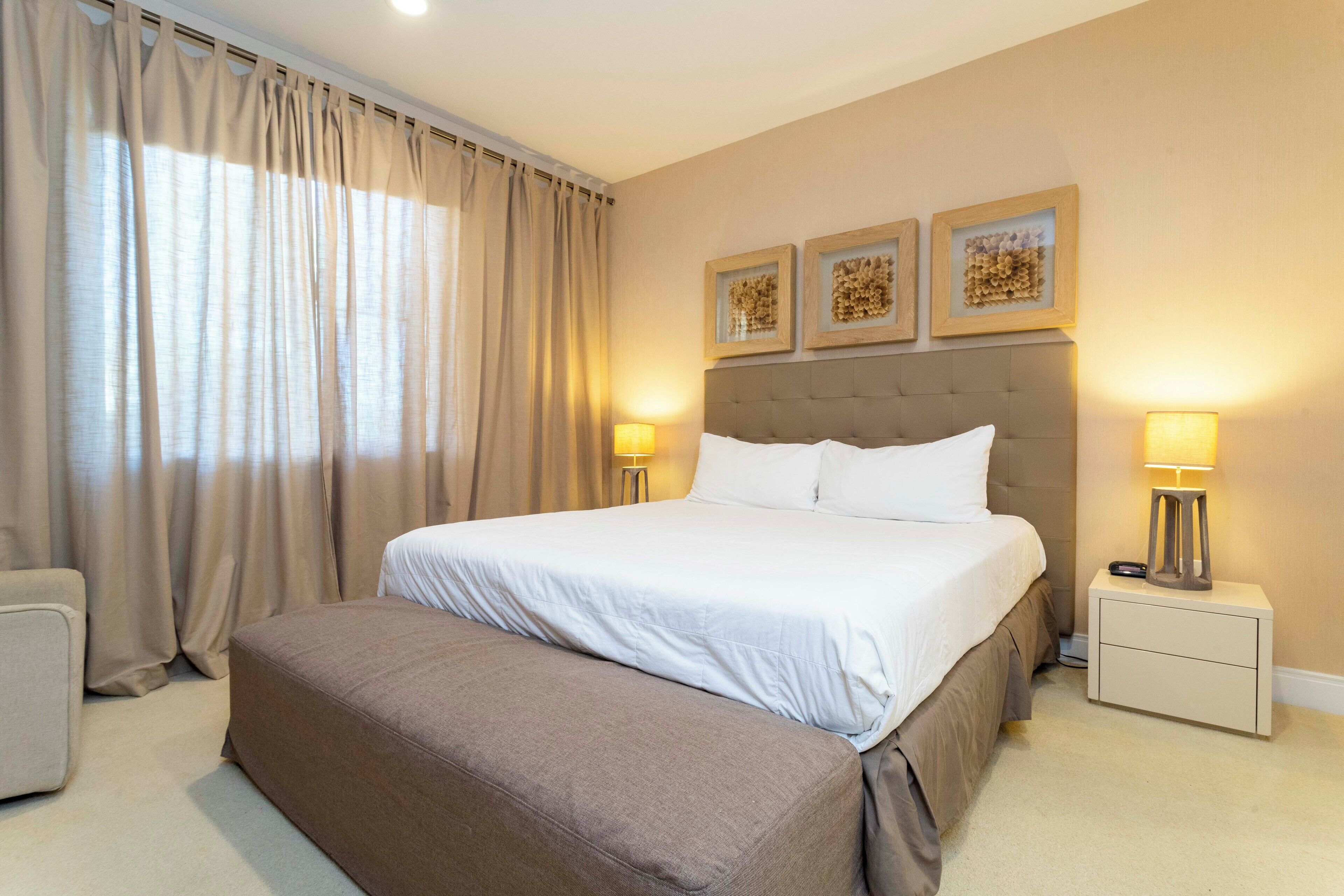 A neatly arranged bedroom with a large bed, two bedside tables with lamps, a small sofa, and three framed wall art pieces.